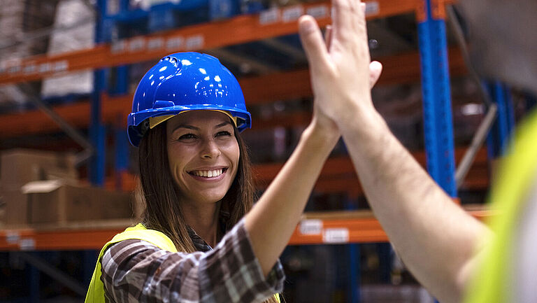 In einem Lager sieht man eine lächelnde Frau mit Schutzhelm und Warnweste beim High Five mit einem Mann.